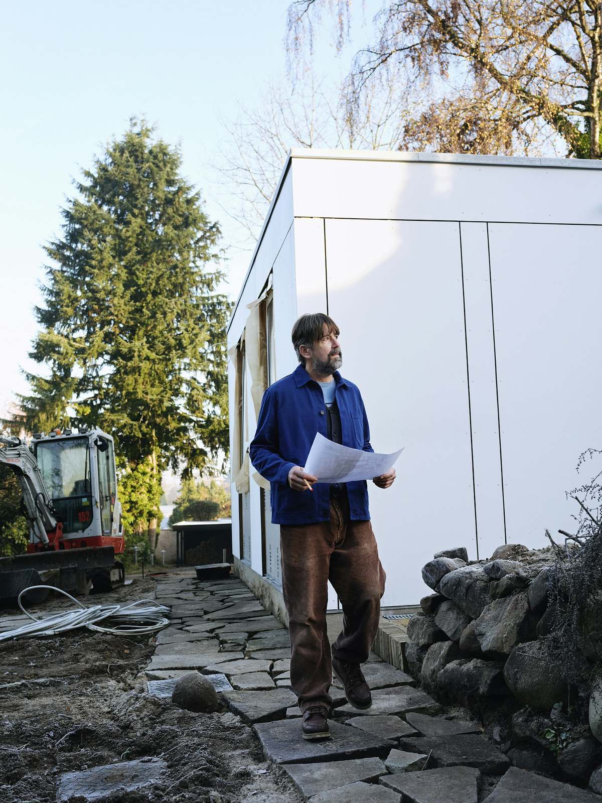 Man met plannen in de hand op een werf naast een wit gebouw en een graafmachine